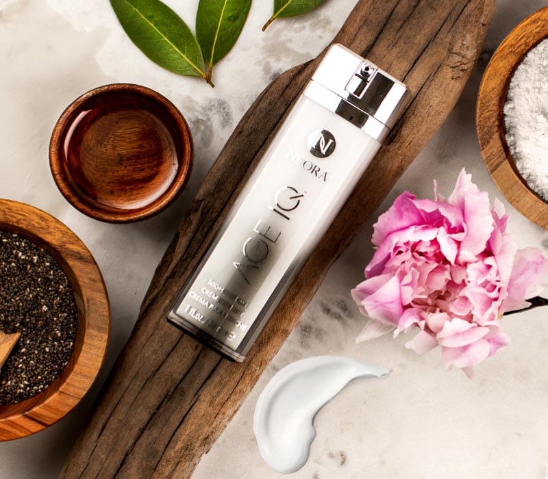 Neora’s best-selling Age IQ® Night Cream laying on a wooden plant next to a leaf, flower, a cup of oil and a bowl of seeds